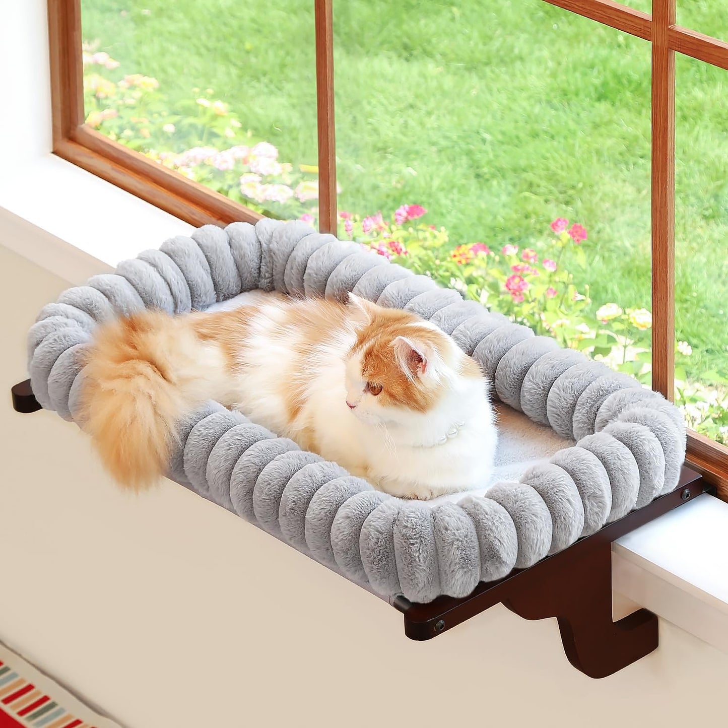 Cat lounging on a fluffy pet bed attached to a window ledge with a view of greenery outside.